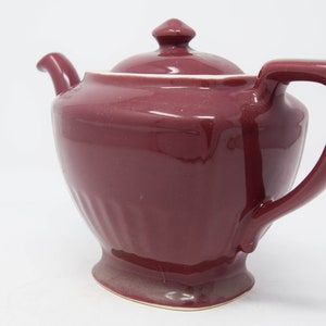 May include: A maroon ceramic teapot with a lid and a handle. The teapot has a slightly rounded body and a flared base.