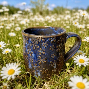 May include: A ceramic mug with a deep blue and brown glaze featuring a repeating embossed flower pattern. The mug has a sturdy handle and a smooth, glossy interior finish, showcasing a dark blue hue that contrasts with the textured exterior.