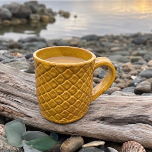 May include: A mustard yellow ceramic mug with a textured, repeating diamond pattern across its exterior surface. The mug features a curved handle on the right side and is filled with a light brown liquid, likely coffee or tea, visible at the top.