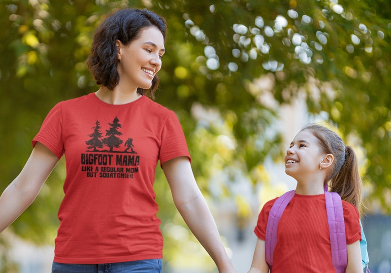 A red t-shirt with black text that reads 'BIGFOOT MAMA LIKE A REGULAR MOM BUT SQUATCHIER' and a graphic of a Bigfoot walking in front of two trees.