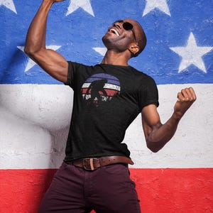 Puede incluir: Camiseta negra con un gráfico de Bigfoot en rojo, blanco y azul, usada con pantalones burdeos y un cinturón marrón. El fondo presenta un diseño de bandera americana pintada con estrellas blancas sobre un campo azul.