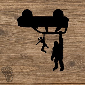 May include: A black vinyl decal featuring a silhouette of an overturned off-road vehicle with a person and a large hairy creature hanging from the chassis, accompanied by a small footprint icon in the lower left corner of the design.