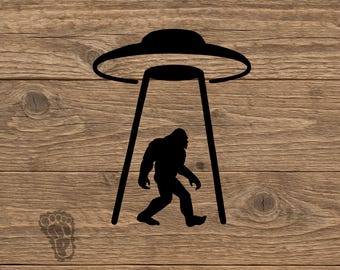 May include: A black silhouette graphic featuring a Bigfoot figure walking beneath a hovering UFO with a tractor beam. A small, faint footprint icon is positioned in the lower left corner of the design, which is presented on a wood grain background.