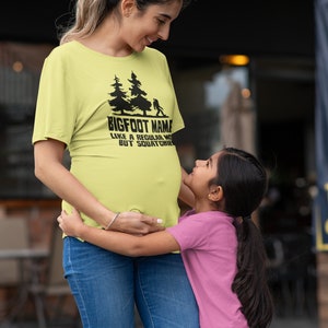 A pregnant woman wearing a yellow t-shirt with the text 'BIGFOOT NAME LIKE A REGULAR MOM BUT SQUATCHIER' and a young girl wearing a pink t-shirt hugging her belly.