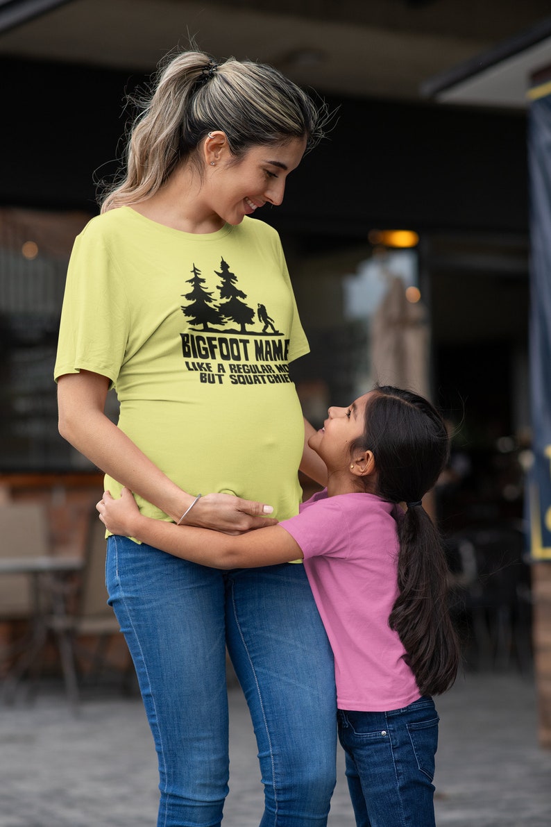 A pregnant woman wearing a yellow t-shirt with the text 'BIGFOOT NAME LIKE A REGULAR MOM BUT SQUATCHIER' and a young girl wearing a pink t-shirt hugging her belly.