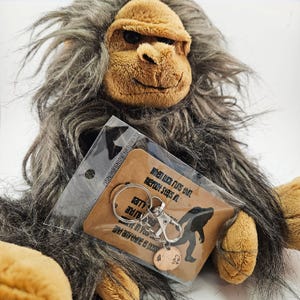 May include: A plush Bigfoot doll with brown fur and a smiling face. A keychain with a Bigfoot silhouette and the text "When luck runs thin, Bigfoot steps in" is in front of the doll.