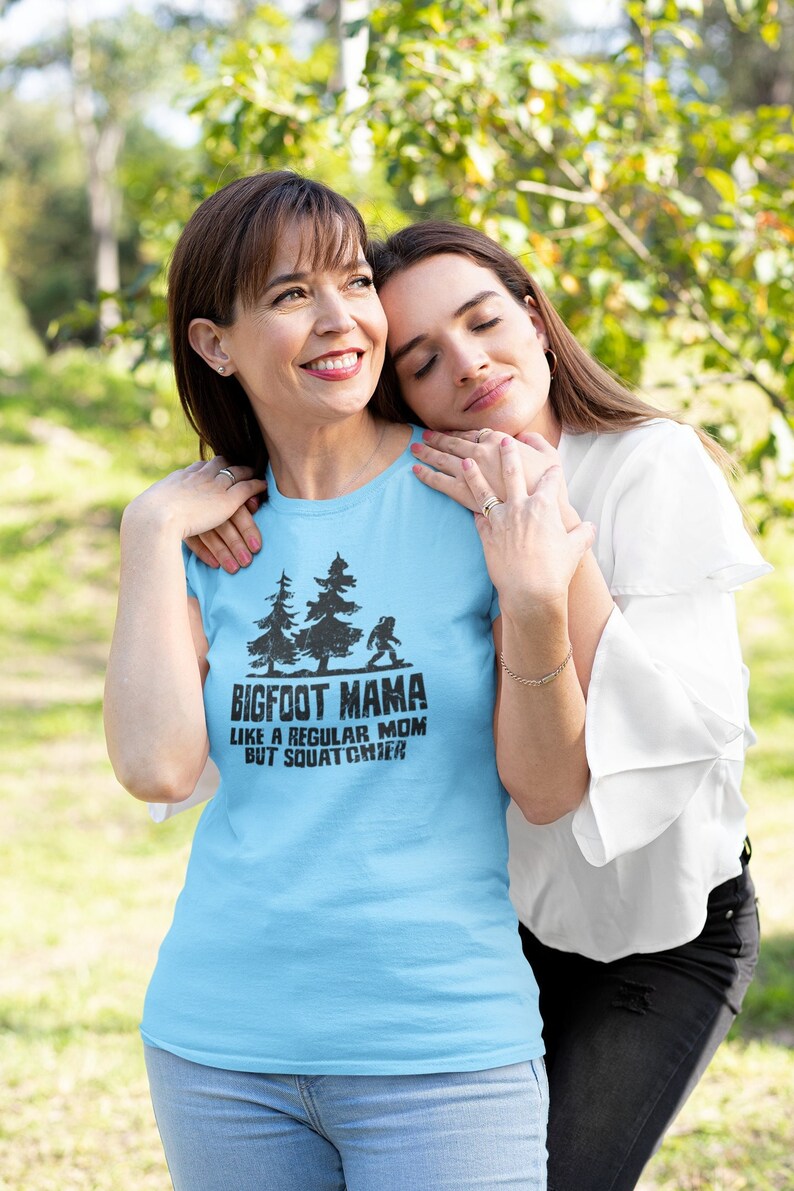 t-shirt with a black graphic of two trees and a Bigfoot. The text on the shirt reads 'Bigfoot Mama Like a Regular Mom But Squatchier.'