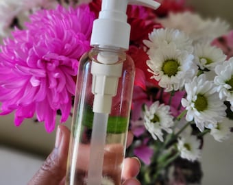 May include: A clear glass bottle with a white pump dispenser filled with a light yellow liquid. The bottle is being held by a hand in front of a bouquet of pink and white flowers.