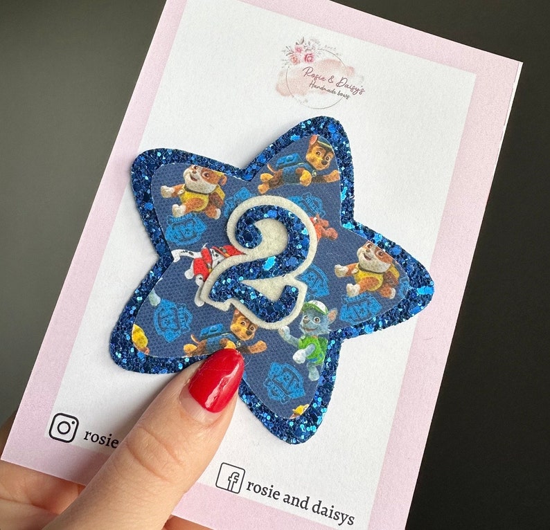 May include: Blue glitter star-shaped applique with a white felt number 2 in the centre. The star is covered in a Paw Patrol print fabric featuring the characters Chase, Marshall, Skye, and Rubble.
