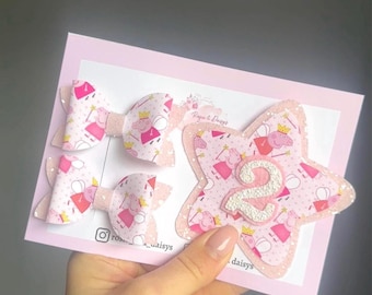 May include: Pink hair bows and a star-shaped decoration with the number 2. The bows and star feature a repeating pattern of a cartoon character. The star is outlined in glitter and has a pink and white background.