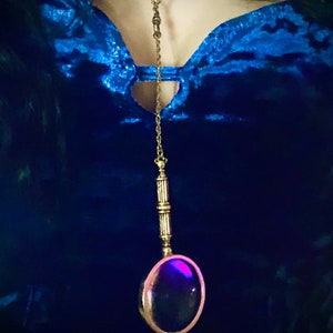 May include: A gold chain necklace with a magnifying glass pendant. The magnifying glass is round and has a dark purple lens. The necklace is worn over a blue velvet top.