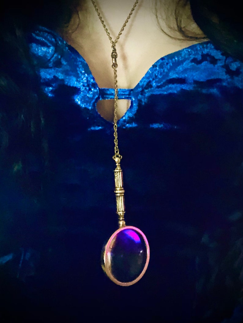 May include: A gold chain necklace with a magnifying glass pendant. The magnifying glass is round and has a dark purple lens. The necklace is worn over a blue velvet top.