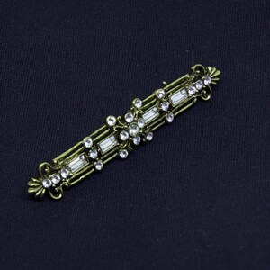 May include: A rectangular bar brooch in an antique brass finish featuring a central cluster of clear round rhinestones, accented by smaller round stones and rectangular baguette crystals arranged in a symmetrical, openwork metal frame design.