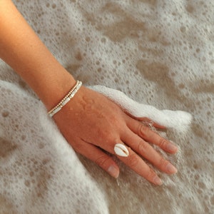 Op de afbeelding: Een hand met een ring gemaakt van een witte schelp op een zanderig strand met wit schuim.