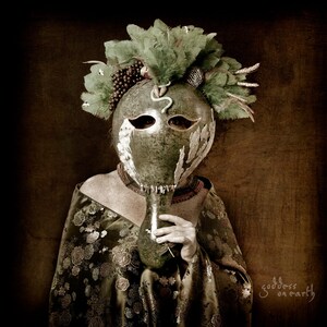May include: A woman wearing a green and white gourd mask with feathers and beads. The mask is held to her face with a hand. The background is a dark brown.