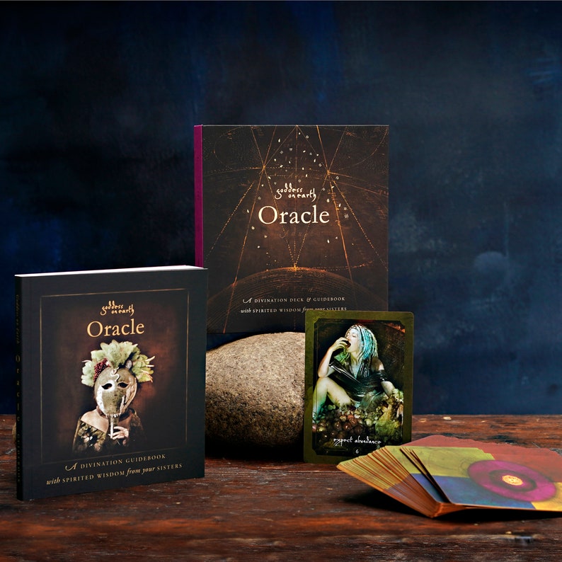 Pu&ograve; includere: Un set di carte oracle con un libro guida. Il libro guida ha uno sfondo scuro con una donna che indossa una maschera e una corona di fiori. Le carte hanno uno sfondo dorato con una donna seduta su uno scoglio. Il testo sul libro guida recita "Dea sulla Terra Oracle" e "Una guida alla divinazione con saggezza ispirata dalle tue sorelle". Il testo sulle carte recita "Aspettati abbondanza".