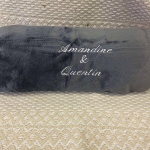 Peut inclure: Un coussin en peluche gris foncé avec une broderie blanche qui dit "Amandine & Quentin".