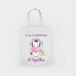 Peut inclure: Sac en toile blanc avec une licorne violette lisant un livre et le texte "Le sac de bibliotheque d'Agathe".