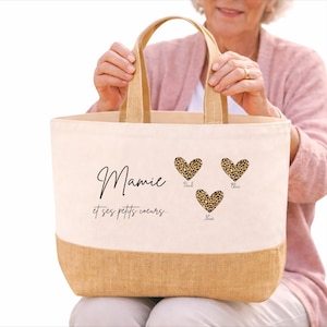 K&ouml;nnte beinhalten: Eine Canvas-Tragetasche mit einem Boden und Griffen aus Naturjute. Die Tasche zeigt den Text "Mamie et ses petits coeurs..." und drei Herzen mit Leopardenmuster und Namen. Die Tasche wird von einer Person gehalten.