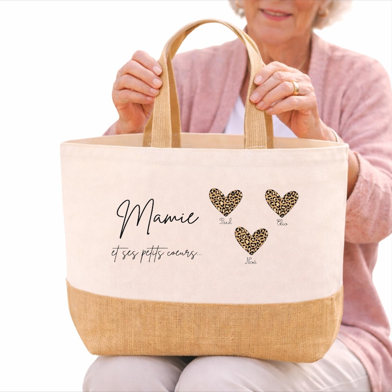 K&ouml;nnte beinhalten: Eine Canvas-Tragetasche mit einem Boden und Griffen aus Naturjute. Die Tasche zeigt den Text "Mamie et ses petits coeurs..." und drei Herzen mit Leopardenmuster und Namen. Die Tasche wird von einer Person gehalten.