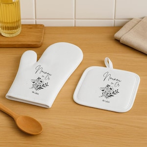 May include: White oven mitt and matching square pot holder set. Both items feature a floral design and the text "Nounou en Or" in black script. The pot holder has a loop for hanging.