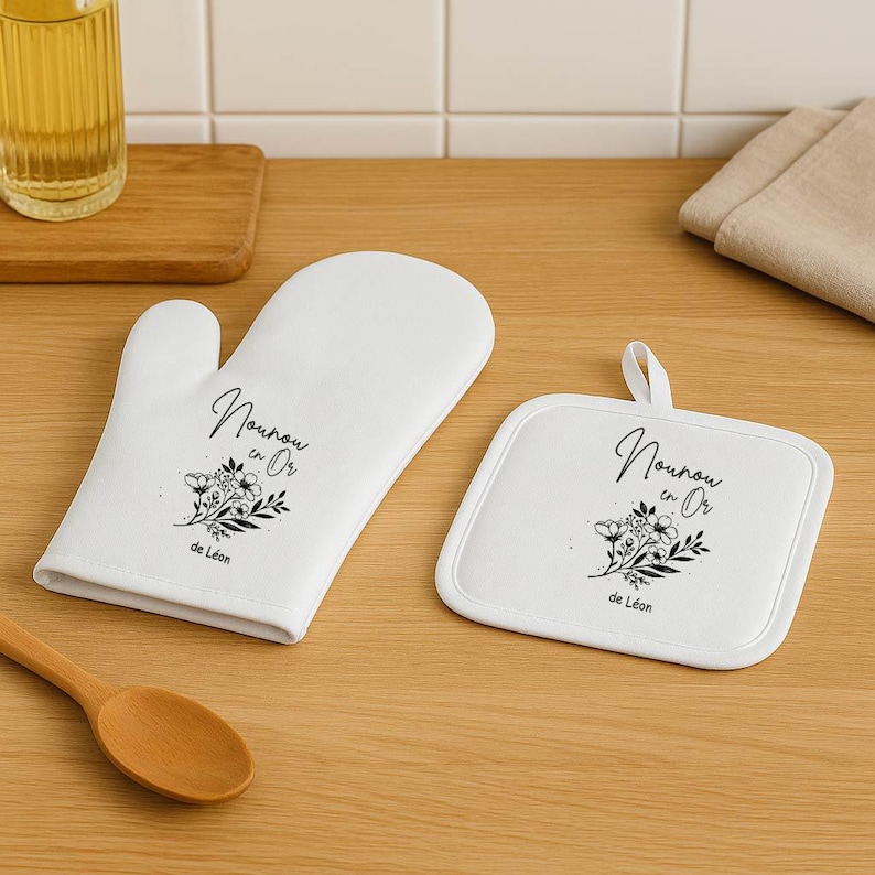 May include: White oven mitt and matching square pot holder set. Both items feature a floral design and the text "Nounou en Or" in black script. The pot holder has a loop for hanging.
