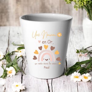 Peut inclure: Mug en céramique blanche avec un motif arc-en-ciel et cœur. Le mug porte l'inscription dorée "Une Nounou en Or" et "ça reste dans le coeur Paul". Décoré d'un arc-en-ciel et de cœurs.