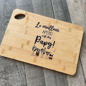 Peut inclure: Une planche à découper en bois avec le texte "Le meilleur APERO c'est chez Papy!" et une rangée de cinq verres à cocktail.