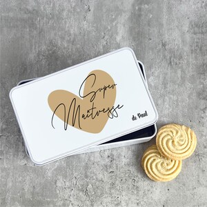 Peut inclure: Boîte en métal blanc avec un cœur doré et le texte "Super Maitresse de Paul". La boîte est ouverte et il y a deux biscuits en forme de spirale à côté.