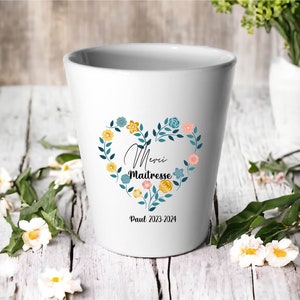 Peut inclure: Un pot en céramique blanc avec un motif floral en forme de cœur. Le cœur est entouré d'une couronne de feuilles vertes et de fleurs colorées. Le texte "Merci Maitresse" est à l'intérieur du cœur, et le texte "Paul 2023-2024" est en dessous du cœur.
