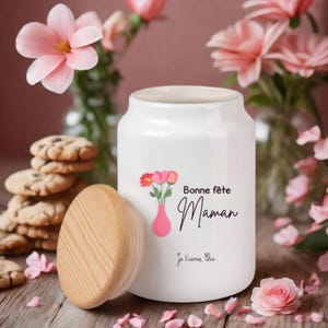 Peut inclure: Un pot en céramique blanc avec un couvercle en bois, décoré d'un motif floral et du texte "Bonne fête Maman" en écriture noire. Le pot est entouré de fleurs roses et de biscuits.