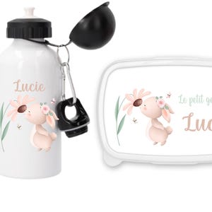 Peut inclure: Ensemble gourde et boîte à lunch blancs avec un lapin de dessin animé et une fleur. Le nom "Lucie" est inscrit sur les deux articles. La boîte à lunch porte l'inscription "Le petit goûter de Lucie".