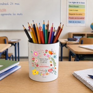 Peut inclure: Un pot à crayons blanc rempli de crayons de couleur sur un bureau en bois dans une salle de classe. Le pot est décoré d'un motif floral et de texte en français. Un cahier et d'autres fournitures scolaires sont également sur le bureau.