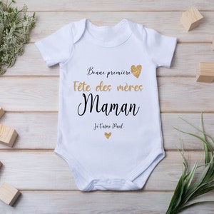 Peut inclure: Un body blanc pour bébé avec le texte "Bonne premiere Fete des meres Maman Je t'aime Paul" en noir et paillettes dorées. Le body est sur un fond en bois avec de petits blocs en bois et de la verdure.
