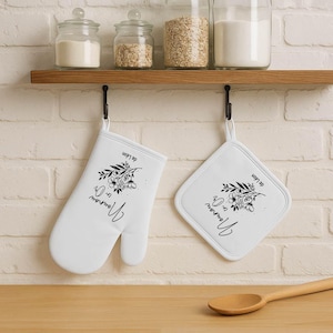 May include: White oven mitt and pot holder set with black floral design and script text. The text reads "Nounou en Or" and "de L&eacute;on". The set hangs from a black hook below a wooden shelf.