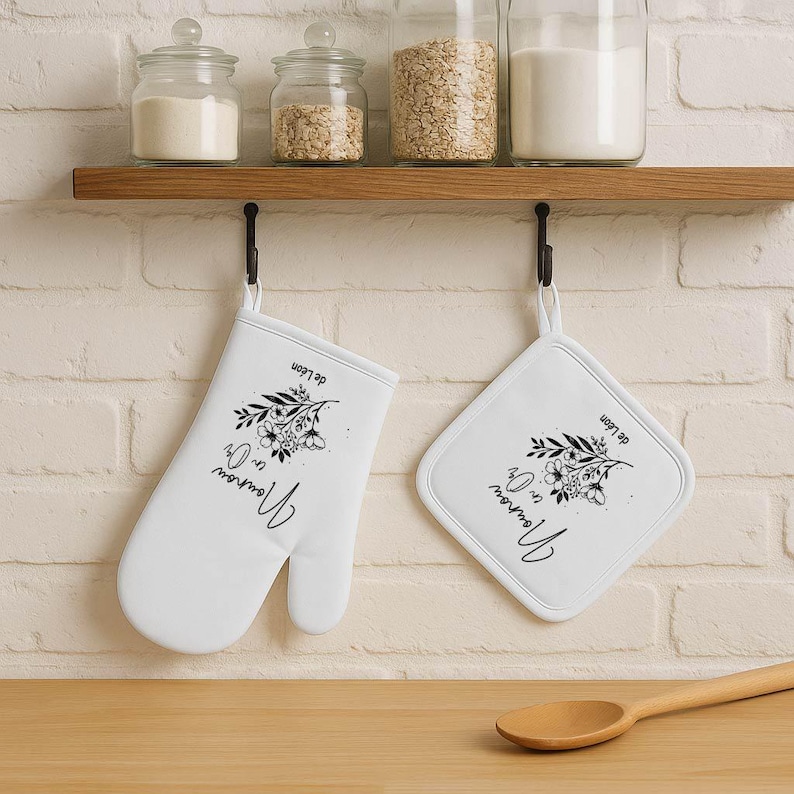 May include: White oven mitt and pot holder set with black floral design and script text. The text reads "Nounou en Or" and "de L&eacute;on". The set hangs from a black hook below a wooden shelf.