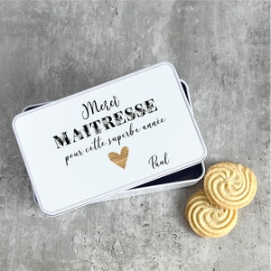 Peut inclure: Boîte en métal blanc avec un design noir et or. La boîte porte le texte "Merci Maitresse pour cette superbe année" et un cœur doré. Le nom "Paul" est écrit sous le cœur. La boîte est ouverte et il y a deux biscuits sur une surface grise.