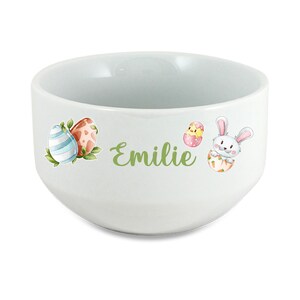 Peut inclure: Bol en céramique blanche avec un motif de Pâques vert représentant un lapin, un poussin et des œufs. Le bol est personnalisé avec le nom "Emilie".