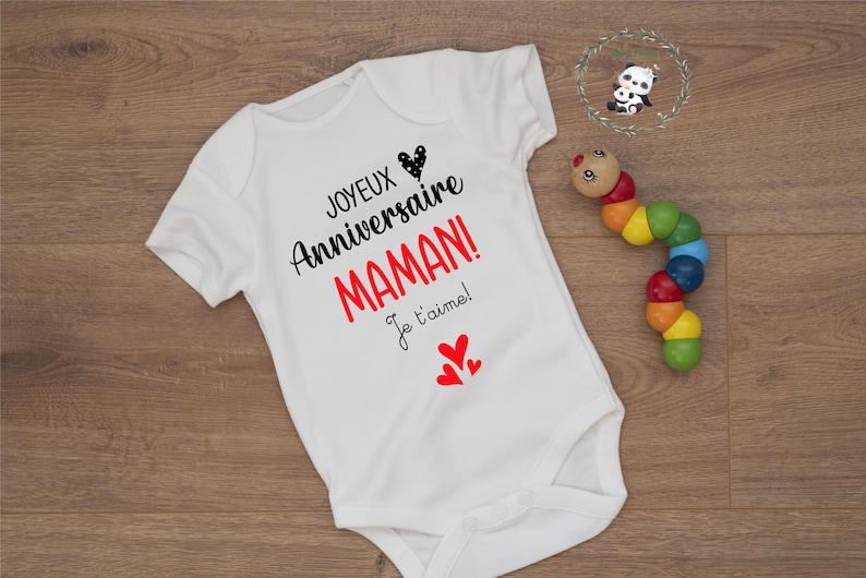 May include: White baby onesie with black text that reads "Joyeux Anniversaire Maman! Je t'aime!" with red hearts.