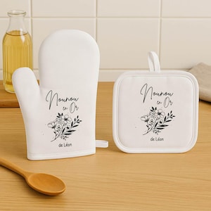 May include: White oven mitt and square pot holder set. Each item features a black floral design and the text "Nounou en Or de L&eacute;on". The oven mitt is on the left, and the pot holder is on the right.