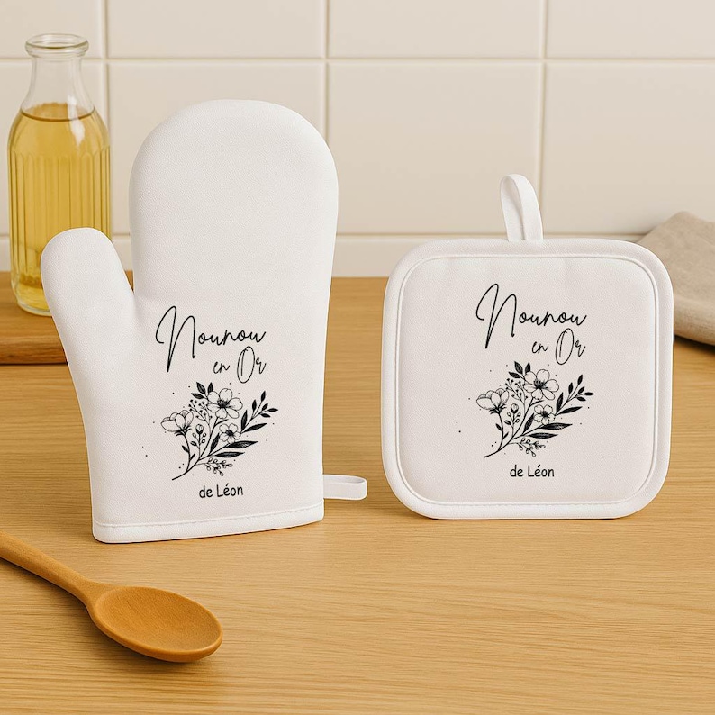 May include: White oven mitt and square pot holder set. Each item features a black floral design and the text "Nounou en Or de L&eacute;on". The oven mitt is on the left, and the pot holder is on the right.