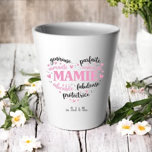 Peut inclure: Pot de fleurs en céramique blanche avec le mot "MAMIE" en rose, entouré de mots comme "généreuse" et "adorable" en forme de cœur. Le pot a un design simple et convient aux petites plantes.