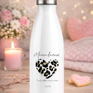 Peut inclure: Une gourde isotherme blanche avec un couvercle et une base argentés. La gourde présente un motif de cœur léopard noir et or avec les mots "Mamie amour" et d'autres textes. Une bougie et des fleurs sont en arrière-plan.