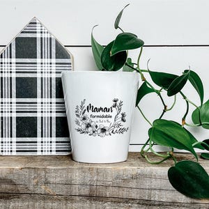 Peut inclure: Pot en céramique blanche avec un motif floral noir et le texte "Maman formidable" en lettres noires. Le pot est rempli d'une plante verte avec des vignes retombantes.
