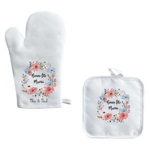Peut inclure: Ensemble de maniques et de dessous de plat blancs avec un motif de couronne florale. La couronne est ornée de fleurs roses et bleues et du texte "Bonne fête Mamie Clio & Paul".