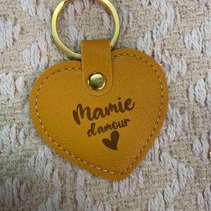 May include: A yellow heart-shaped leather keychain with a gold ring and a gold snap closure. The keychain has the text "Mamie d'amour" and a small heart engraved on it.
