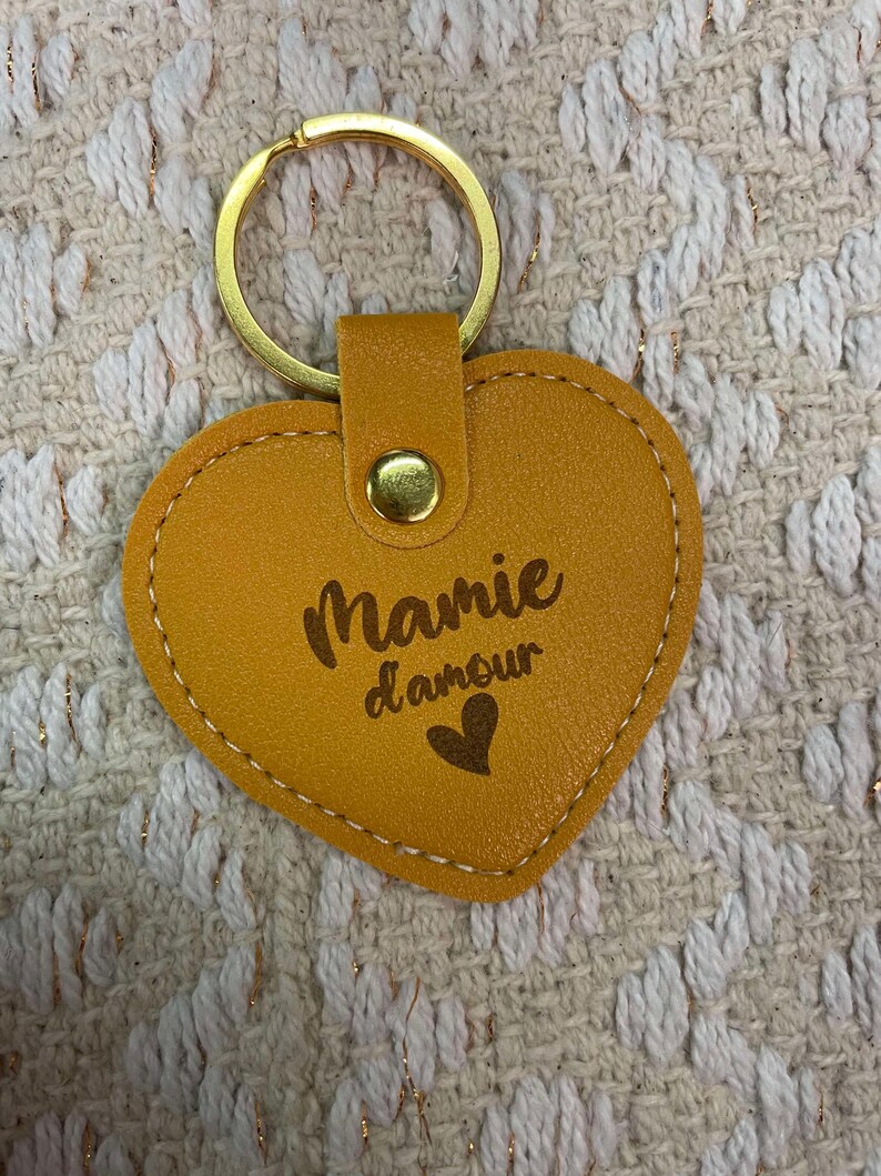 May include: A yellow heart-shaped leather keychain with a gold ring and a gold snap closure. The keychain has the text "Mamie d'amour" and a small heart engraved on it.