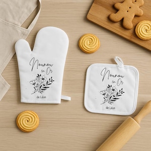 May include: White oven mitt and pot holder set with floral design and text. The text reads "Nounou en Or" and "de L&eacute;on" in black script. The set is accompanied by cookies and a rolling pin.