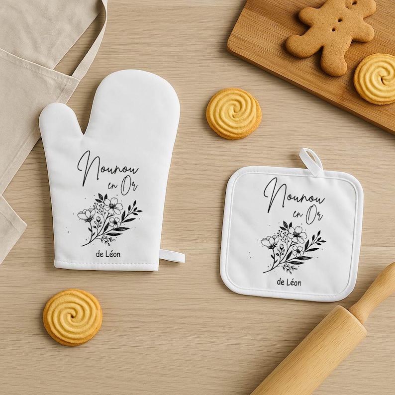 May include: White oven mitt and pot holder set with floral design and text. The text reads "Nounou en Or" and "de L&eacute;on" in black script. The set is accompanied by cookies and a rolling pin.