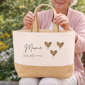 K&ouml;nnte beinhalten: Beige Tragetasche mit einem Boden und Griffen aus Naturjute. Auf der Tasche steht das Wort "Mamie" in Schreibschrift, darunter "et ses petits coeurs". Drei Leopardenherzen sind auf der Tasche aufgedruckt, jedes mit einem Namen.